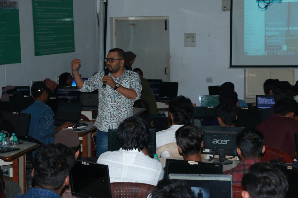 Naffys Mir speaking to students in a computer lab, holding a microphone.